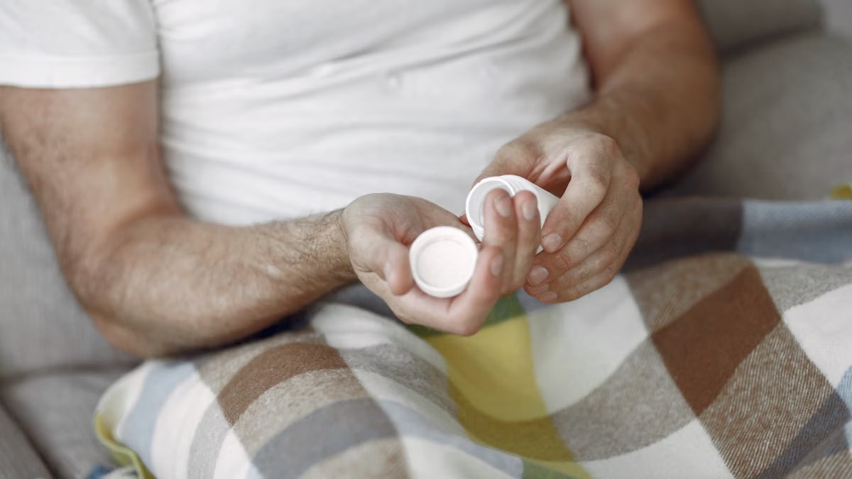 Adult man sitting on sofa, holding pills, wrapped in cozy blanket, relaxed setting.